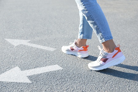 Person wearing jeans and sneakers standing on asphalt road, two white arrows pointing in different directions