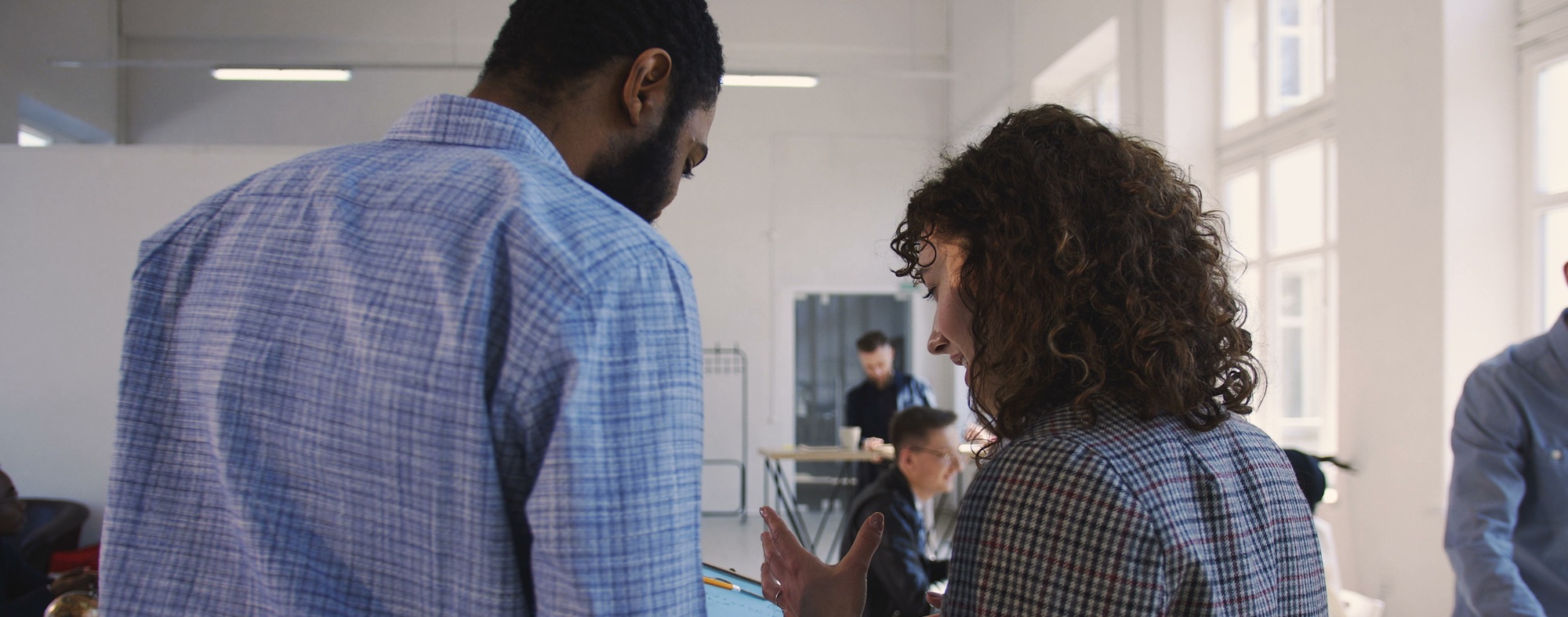 Man and woman in office standing with backs turned toward camera, having a conversation and pointing at a laptop.