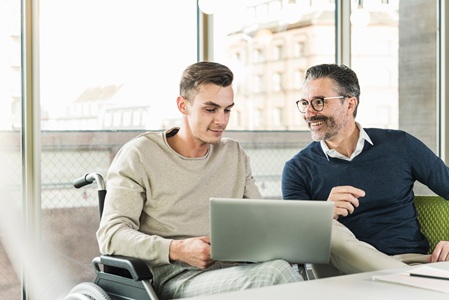 A man with glasses and facial hair sitting by a desk with a younger man in a wheelchair, engaged in conversation and looking at a laptop.